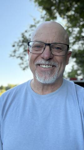 Smiling man with glasses and a white beard, outdoors with trees in the background.